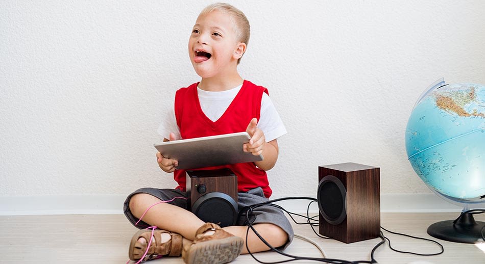Boy with down syndrome sits crossed leg on the floor, playing music from an Ipad. 