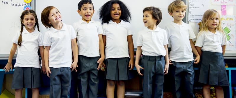 A group of primary school children in white polo shirts and grey skirts and trousers stand in front of their classroom board smiling at the camera.